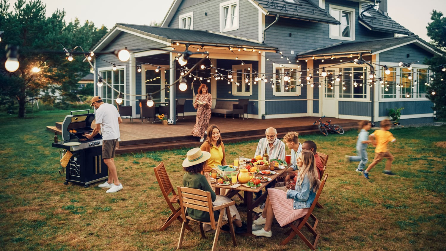 A family having dinner in their backyard with a grill, outdoor table, string lights, and an excellent speaker system as part of their whole home audio.